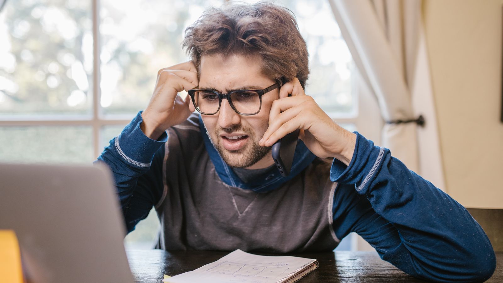 Man in glasses using a laptop and smartphone at home, appearing confused