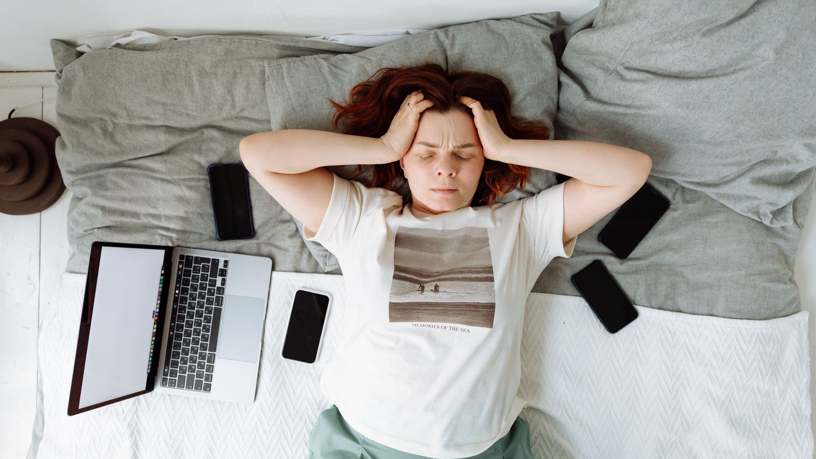 Distressed woman lying in bed surrounded by digital devices, showing signs of stress